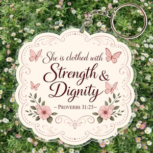 May include: A clear acrylic keychain with a scalloped edge, featuring the text "She is clothed with Strength & Dignity - PROVERBS 31:25". The design includes pink butterflies and floral accents, attached to a silver keyring.