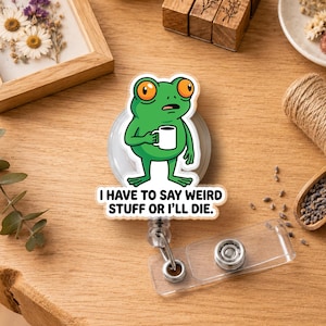 May include: A circular badge reel featuring a green frog illustration holding a white coffee mug. Below the frog, the text reads: I HAVE TO SAY WEIRD STUFF OR I'LL DIE. The badge reel has a clear plastic strap with a metal snap button attachment.
