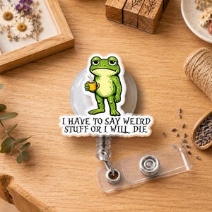 May include: A circular badge reel featuring a green frog illustration holding a yellow mug. Below the frog, a white speech bubble shape contains the black text: I HAVE TO SAY WEIRD STUFF OR I WILL DIE. The badge reel has a clear plastic strap.
