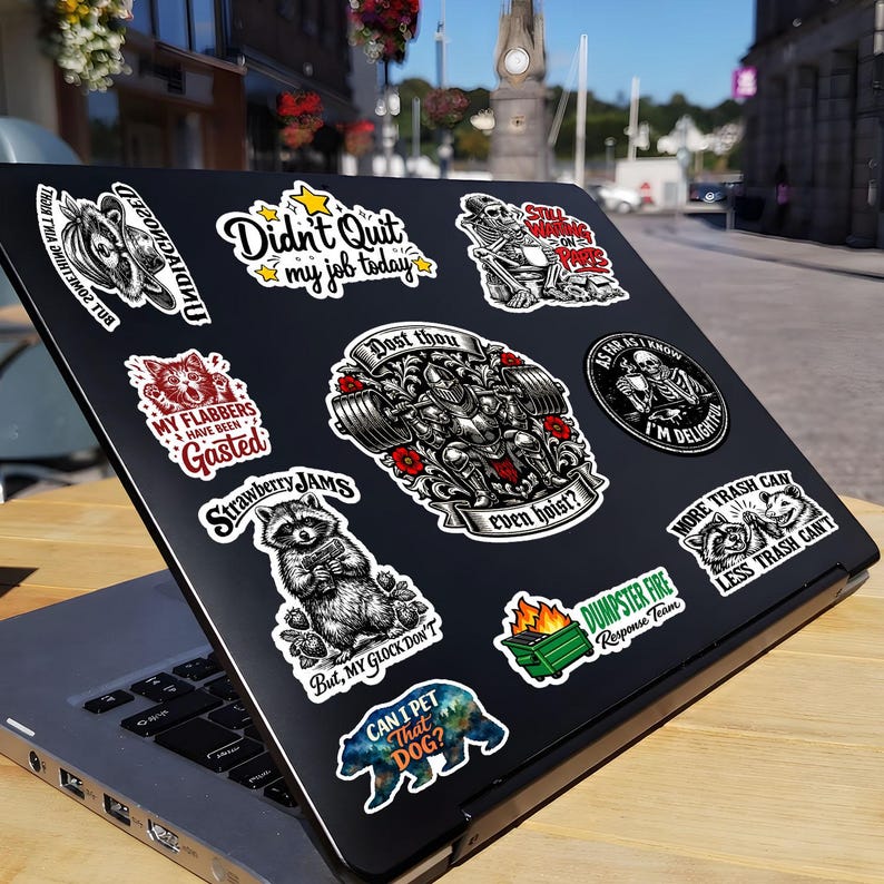 Puede incluir: Un ordenador port&aacute;til negro cubierto con varias pegatinas. Las pegatinas incluyen frases como "Didn't Quit my job today" y "Can I pet that dog?". Las pegatinas presentan ilustraciones de un mapache, un oso y un esqueleto.