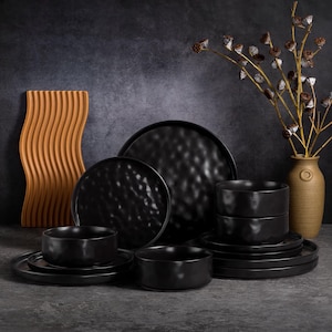 May include: A collection of black ceramic dinnerware, including plates and bowls, arranged on a dark surface. A decorative, wavy, orange-brown piece and a vase with dried flowers are also present, creating a stylish table setting.