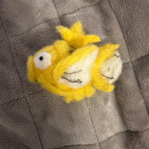 May include: A handmade yellow felt fish with white accents and black details. The fish has a crown-like fin and a round eye. The felt fish is laying on a gray, textured surface.