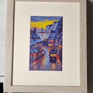 May include: Framed art print depicting a European coastal town at dusk. The artwork features a street with a trolley, buildings with blue roofs, and a lighthouse in the distance. The sky is a mix of yellow and orange hues, and the print is framed in a light gray wooden frame.