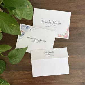 May include: White envelopes with floral and botanical designs. The envelopes have the names "Mr and Mrs John Doe" and "The Smiths" printed on them, along with address information. The envelopes are arranged on a dark wood surface.