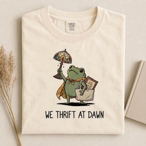 May include: A cream-colored t-shirt featuring a graphic of a green frog wearing a patterned scarf, holding a stained-glass lamp, and carrying a tote bag with a picture frame inside. The text below the frog reads, WE THRIFT AT DAWN.