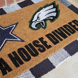 May include: A rectangular tan coir doormat featuring a navy blue star on the left, a green and white eagle logo on the top right, and the black text A HOUSE DIVIDED printed across the bottom right, all layered over a black and white buffalo plaid rug.