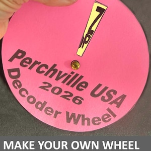 Op de afbeelding: Een roze decoderwiel met de tekst "Perchville USA 2026 Decoder Wheel". Het wiel heeft een gele uitroepteken en een kleine goudkleurige edelsteen. De tekst "Make Your Own Wheel and Decipher Messages!" staat onderaan.