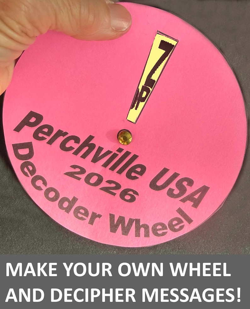 Op de afbeelding: Een roze decoderwiel met de tekst "Perchville USA 2026 Decoder Wheel". Het wiel heeft een gele uitroepteken en een kleine goudkleurige edelsteen. De tekst "Make Your Own Wheel and Decipher Messages!" staat onderaan.