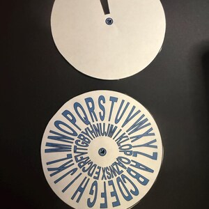 Op de afbeelding: Twee witte cirkelvormige schijven op een zwarte achtergrond. De bovenste schijf heeft een uitroepteken uitsparing. De onderste schijf toont het alfabet in blauw, in een cirkel gerangschikt. Beide schijven hebben een kleine blauwe bevestiging in het midden.