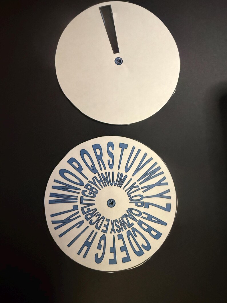 Op de afbeelding: Twee witte cirkelvormige schijven op een zwarte achtergrond. De bovenste schijf heeft een uitroepteken uitsparing. De onderste schijf toont het alfabet in blauw, in een cirkel gerangschikt. Beide schijven hebben een kleine blauwe bevestiging in het midden.