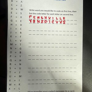 Op de afbeelding: Een wit papier met de titel "Perchville USA Decoder Wheel - 2026" en instructies voor het maken van een gecodeerd bericht. Het woord "PERCHVILLE" is met rode inkt geschreven, met de gecodeerde vorm eronder. Er is ook een codeersleutel aanwezig.