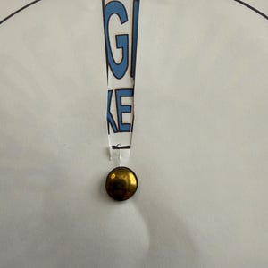 Op de afbeelding: Een wit papier met het woord "GIVE" uitgesneden, waardoor blauwe letters zichtbaar worden. Onder de uitgesneden tekst is een goudkleurige, ronde knoop bevestigd. Het papier heeft een zwarte cirkelvormige omtrek.