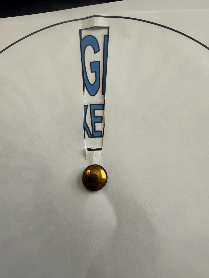 Op de afbeelding: Een wit papier met het woord "GIVE" uitgesneden, waardoor blauwe letters zichtbaar worden. Onder de uitgesneden tekst is een goudkleurige, ronde knoop bevestigd. Het papier heeft een zwarte cirkelvormige omtrek.