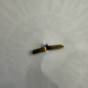 Op de afbeelding: Een klein, messingkleurig metalen object met een puntig uiteinde en een kleine blauwe accent. Het object lijkt een speld of een vergelijkbaar bevestigingsmiddel te zijn. De achtergrond is een wit oppervlak met vage tekst.