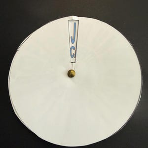 Op de afbeelding: Een wit cirkelvormig object met een messingkleurige knop in het midden. Het object heeft een uitsparing aan de bovenkant met de letters "JC" in blauw. De achtergrond is zwart.
