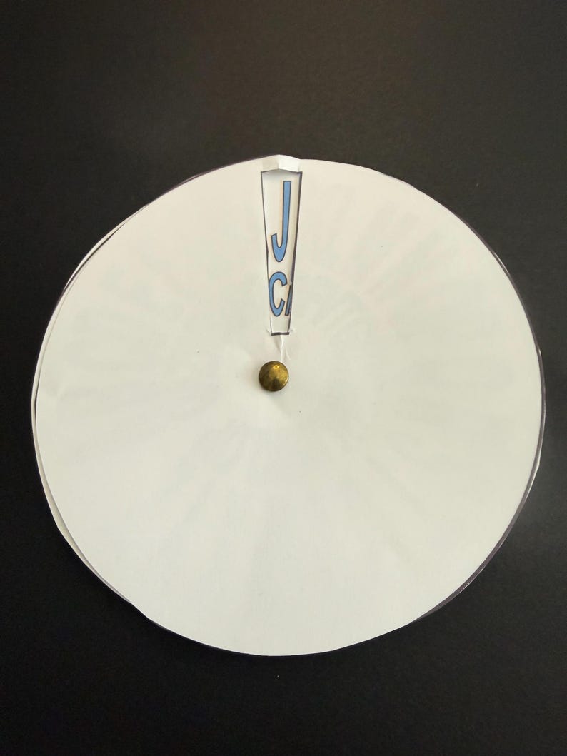 Op de afbeelding: Een wit cirkelvormig object met een messingkleurige knop in het midden. Het object heeft een uitsparing aan de bovenkant met de letters "JC" in blauw. De achtergrond is zwart.
