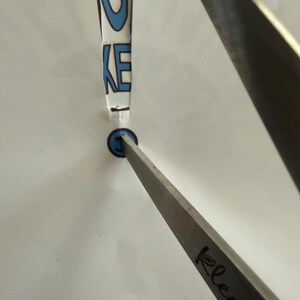 Op de afbeelding: Een doorzichtig object in de vorm van een uitroepteken met de letters "GKE" in blauw. Een blauwe cirkel bevindt zich aan de onderkant. Een zilveren metalen gereedschap is op het object gericht.