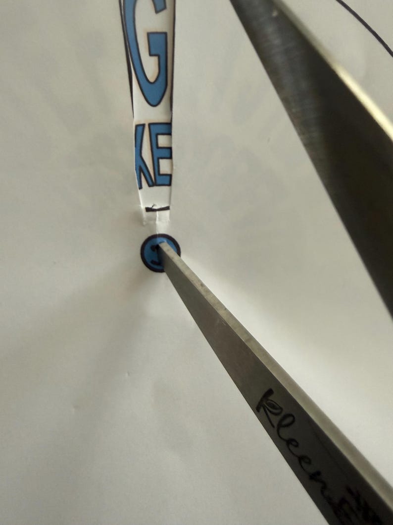 Op de afbeelding: Een doorzichtig object in de vorm van een uitroepteken met de letters "GKE" in blauw. Een blauwe cirkel bevindt zich aan de onderkant. Een zilveren metalen gereedschap is op het object gericht.