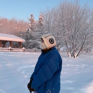 Op de afbeelding: Een persoon draagt een witte gehaakte hoed met een zwart cirkelvormig ontwerp en een bijpassend gezichtsmasker. De persoon draagt een blauwe pufferjas en zwarte handschoenen, staand in een besneeuwd landschap met bomen en een prieel.