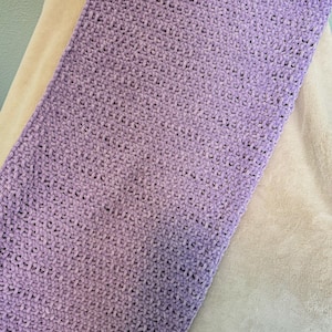 May include: A rectangular, lavender-colored, crocheted blanket. The blanket has a soft, plush texture and is made with a thick yarn. The blanket is resting on a beige surface.