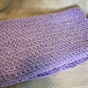 May include: A folded, soft, lavender-colored blanket. The blanket is made of a thick, plush material with a textured, woven pattern. The blanket is folded neatly, showing the layers of the fabric. The overall appearance is cozy and inviting.