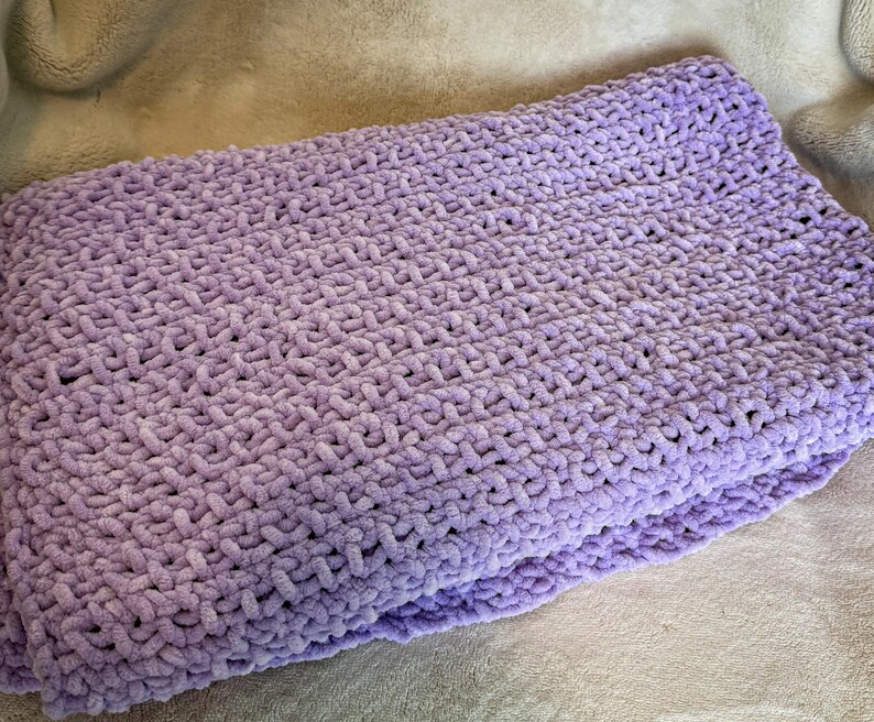 May include: A folded, soft, lavender-colored blanket. The blanket is made of a thick, plush material with a textured, woven pattern. The blanket is folded neatly, showing the layers of the fabric. The overall appearance is cozy and inviting.