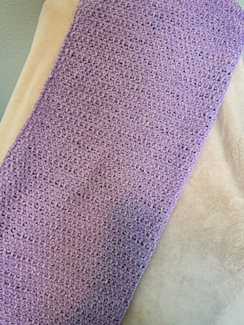 May include: A rectangular, lavender-colored, crocheted blanket. The blanket has a soft, plush texture and is made with a thick yarn. The blanket is resting on a beige surface.