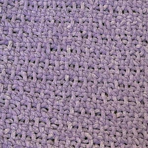 May include: Close-up of a soft, textured, lavender-colored crochet blanket. The stitches are visible, creating a dense, plush surface. The blanket appears to be handmade, with a consistent pattern throughout.