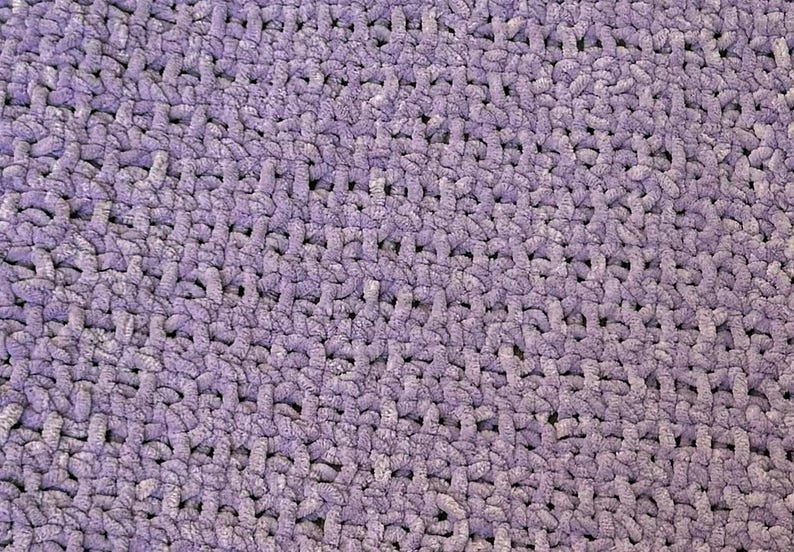 May include: Close-up of a soft, textured, lavender-colored crochet blanket. The stitches are visible, creating a dense, plush surface. The blanket appears to be handmade, with a consistent pattern throughout.