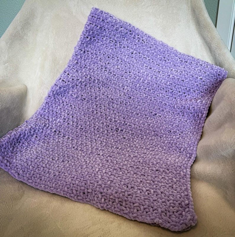 May include: A soft, plush, lavender-colored blanket. The blanket has a gradient effect, transitioning from a darker shade of purple to a lighter shade. The texture appears to be a knitted or crocheted design, creating a cozy and inviting appearance.
