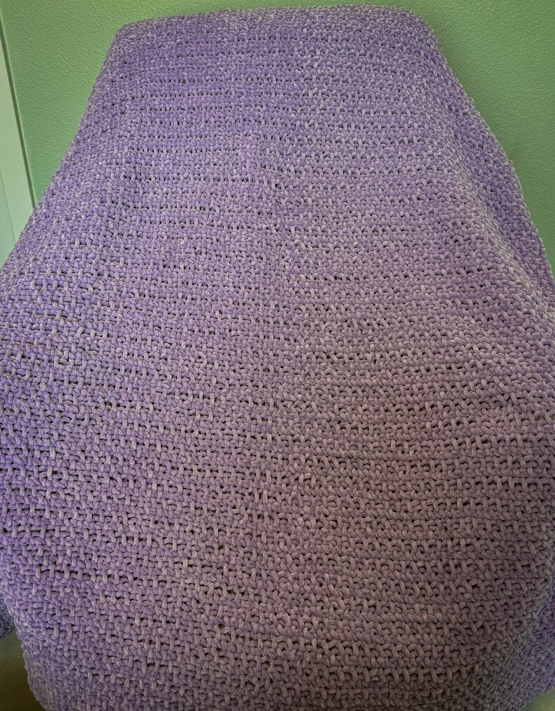 May include: A soft, lavender-colored blanket. The blanket is made of a plush, textured material, likely knitted or crocheted. The color is a light purple, with variations in tone across the surface.