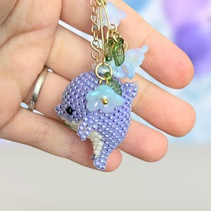 May include: A beaded dolphin charm in shades of purple, white, and green, adorned with delicate blue flower accents. The charm is suspended from a gold chain, creating a whimsical and handcrafted accessory.