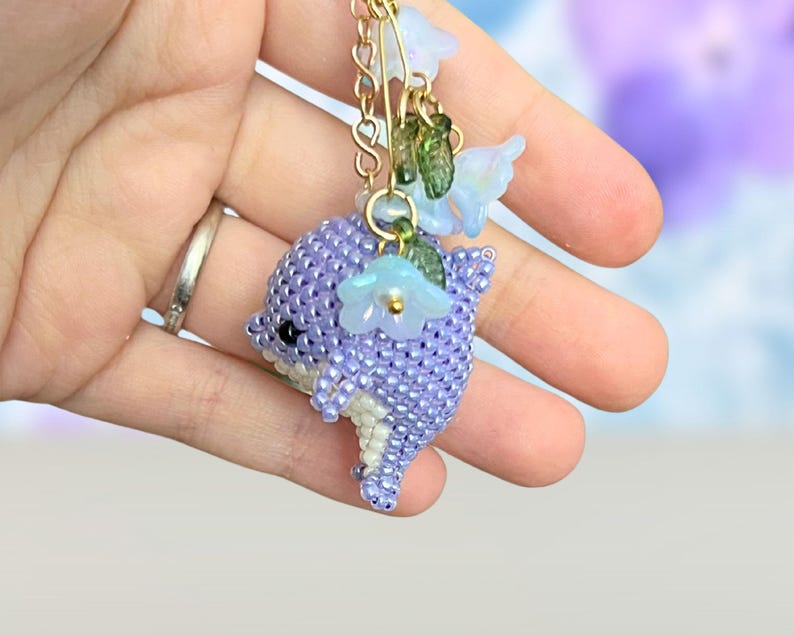 May include: A beaded dolphin charm in shades of purple, white, and green, adorned with delicate blue flower accents. The charm is suspended from a gold chain, creating a whimsical and handcrafted accessory.