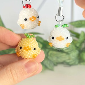 May include: Three small, round, beaded charms shaped like chicks. Two are white with orange beaks and feet, one with a red crest and one with a green leaf on top. The third is yellow with an orange beak and feet and a green leaf on top.