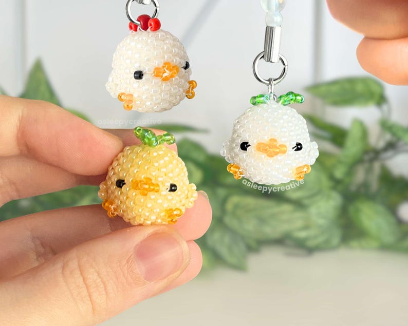 May include: Three small, round, beaded charms shaped like chicks. Two are white with orange beaks and feet, one with a red crest and one with a green leaf on top. The third is yellow with an orange beak and feet and a green leaf on top.