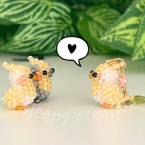 May include: Three small cockatiel figurines crafted from tiny yellow, grey, and peach-colored beads. Two birds stand together on the left, while a third bird with a gold-tone metal loop and a small green leaf charm stands on the right. A white speech bubble with a black heart icon is positioned between them.