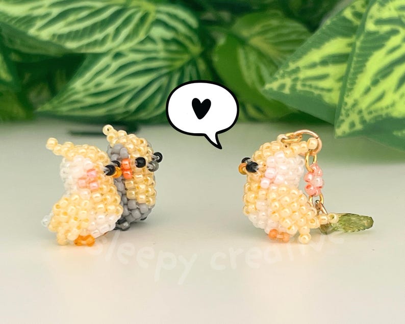 May include: Three small cockatiel figurines crafted from tiny yellow, grey, and peach-colored beads. Two birds stand together on the left, while a third bird with a gold-tone metal loop and a small green leaf charm stands on the right. A white speech bubble with a black heart icon is positioned between them.