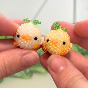 May include: Two small, round, beaded figurines shaped like chicks. One is white with a green leaf on top, and the other is yellow with a green leaf on top. Both have orange beaks, orange feet, and black bead eyes. The text asleepycreative is visible.