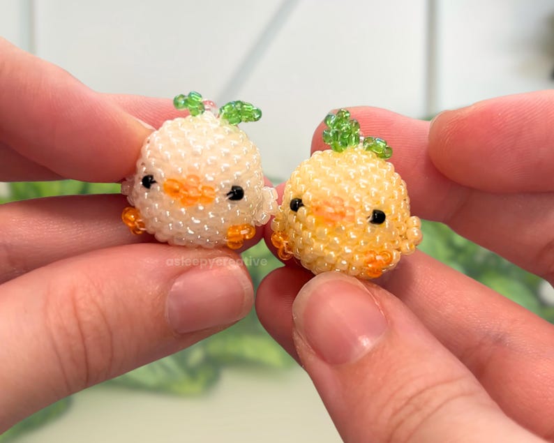 May include: Two small, round, beaded figurines shaped like chicks. One is white with a green leaf on top, and the other is yellow with a green leaf on top. Both have orange beaks, orange feet, and black bead eyes. The text asleepycreative is visible.