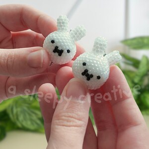 May include: A pair of white beaded bunny earrings. Each earring features a bunny head with black bead eyes and a black bead nose. The earrings are held in a person's hands.