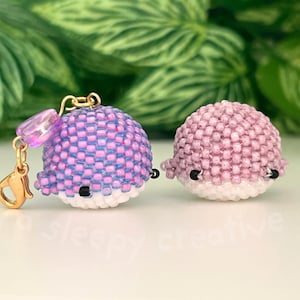 May include: Two beaded whale charms. One is purple and blue, the other is pink. Both have white bellies, black eyes, and small fins. The purple whale has a gold clasp and a purple bead attached.