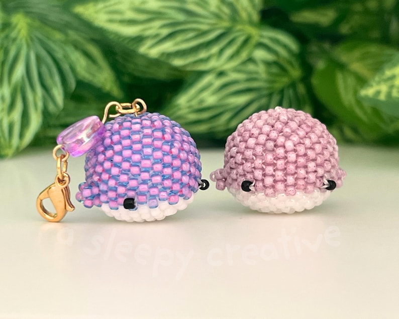 May include: Two beaded whale charms. One is purple and blue, the other is pink. Both have white bellies, black eyes, and small fins. The purple whale has a gold clasp and a purple bead attached.