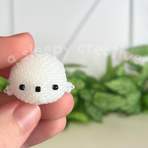 May include: A small, white beaded ghost figurine with black eyes and mouth. The ghost is held in a person's hand, with a blurred green plant background. The figurine has a rounded shape and small, wing-like protrusions on each side.