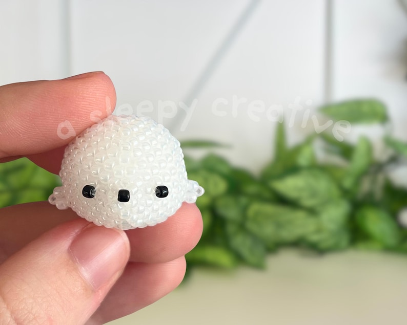 May include: A small, white beaded ghost figurine with black eyes and mouth. The ghost is held in a person's hand, with a blurred green plant background. The figurine has a rounded shape and small, wing-like protrusions on each side.
