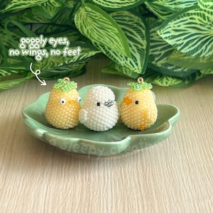 May include: Three beaded bird charms in yellow and white, with green accents, sit on a green, leaf-shaped dish. The charms have googly eyes and are without wings or feet. The dish is on a light-coloured wooden surface, with green foliage in the background.