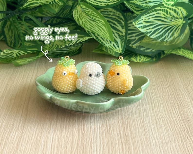 May include: Three beaded bird charms in yellow and white, with green accents, sit on a green, leaf-shaped dish. The charms have googly eyes and are without wings or feet. The dish is on a light-coloured wooden surface, with green foliage in the background.