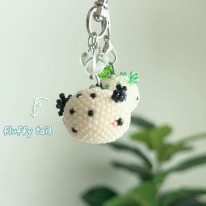 May include: A beaded keychain featuring a round, cream-colored creature with black spots, black appendages, and a small pink accent. The charm hangs from a silver-toned metal clasp with a clear heart-shaped bead. Text reads: fluffy tail.
