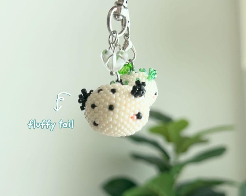 May include: A beaded keychain featuring a round, cream-colored creature with black spots, black appendages, and a small pink accent. The charm hangs from a silver-toned metal clasp with a clear heart-shaped bead. Text reads: fluffy tail.
