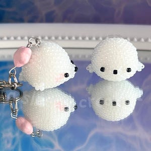 May include: Two white beaded seal charms with black eyes and pink cheeks. One charm is attached to a silver keychain with a pink heart charm. The charms are reflected on a mirrored surface.