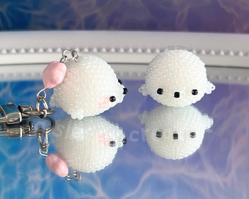 May include: Two white beaded seal charms with black eyes and pink cheeks. One charm is attached to a silver keychain with a pink heart charm. The charms are reflected on a mirrored surface.
